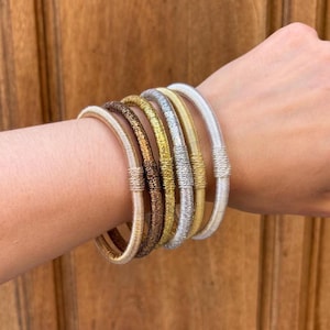 Puede incluir: Una colección de seis pulseras metálicas en varios colores, incluyendo oro, plata y bronce, apiladas en una muñeca. Las pulseras tienen una textura tejida y una forma redondeada.