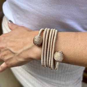 Puede incluir: Un brazalete dorado con un diseño tejido y dos acentos redondos texturizados. El brazalete está envuelto alrededor de la muñeca de una persona.