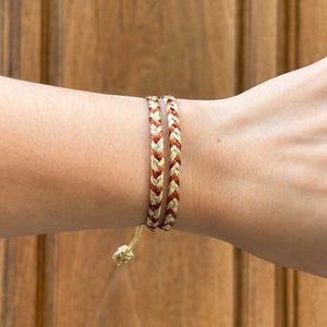 Può includere: Due braccialetti intrecciati in fili dorati e color ruggine sono indossati su un polso. I braccialetti sono fatti di fili intrecciati, creando un aspetto strutturato. I braccialetti sono assicurati con un nodo e un cordino color oro.