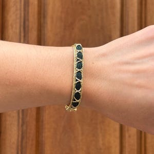 Peut inclure: Un bracelet composé d'un cordon tressé vert foncé central, encadré par deux fines chaînes métalliques dorées torsadées. Le design présente un motif répétitif en forme de X en métal doré qui recouvre le cordon tressé vert foncé.
