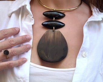 Peut inclure: Un collier ras du cou doré avec trois pendentifs ovales foncés. Le plus grand pendentif est en forme de goutte d'eau. Une bague assortie est également visible.