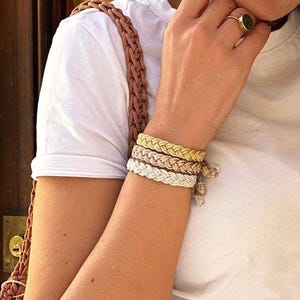 May include: A set of three braided bracelets in metallic gold, rose gold, and silver tones, each featuring a small dangling charm. The bracelets are worn on the wrist alongside a white t-shirt and a brown woven strap, with a gold ring on the finger.
