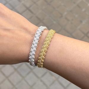 Könnte beinhalten: Zwei geflochtene Armbänder, eines silberfarben und eines goldfarben, werden am Handgelenk getragen. Die Armbänder haben ein strukturiertes, gewebtes Design. Das goldfarbene Armband befindet sich neben dem silberfarbenen Armband.