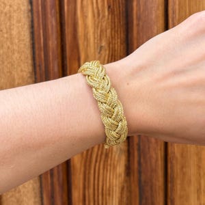 Könnte beinhalten: Ein goldfarbenes geflochtenes Armband. Das Armband besteht aus einem gewebten Material, wodurch ein strukturiertes Aussehen entsteht. Das Armband wird am Handgelenk getragen, mit einem hölzernen Hintergrund.