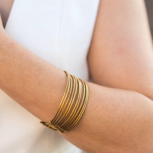 Op de afbeelding: Een goudkleurige armmanchet met meerdere dunne, parallelle banden. De manchet wordt om een arm gedragen, met een witte top zichtbaar op de achtergrond. De sieraden hebben een eenvoudig, elegant ontwerp.