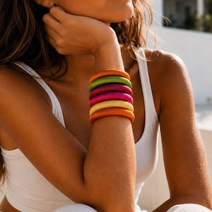 May include: A stack of six cylindrical bangles featuring a smooth, fabric-wrapped texture. The bangles are arranged in a vibrant color sequence consisting of orange, lime green, dark purple, magenta, pale yellow, and a final orange band at the base.