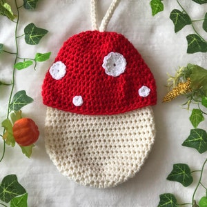 Puede incluir: Una bolsa de ganchillo con forma de hongo con una correa blanca. El hongo es rojo con lunares blancos y detalles de flores blancas.