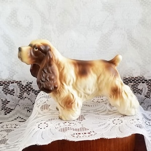 May include: A ceramic figurine of a brown and white cocker spaniel dog. The figurine is standing on a wooden base.