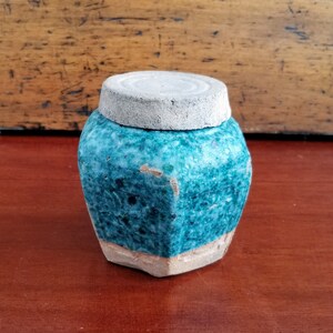 Peut inclure: Un petit pot en céramique de couleur bleu-vert avec un couvercle blanc. Le pot a une surface texturée et une forme légèrement irrégulière.