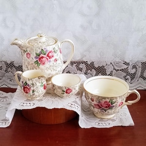 Peut inclure: Un service à thé vintage comprenant une théière, un pot à crème, un sucrier et une tasse. L'ensemble est orné de roses roses, de feuilles vertes et de bordures dorées sur fond crème. Présenté sur une nappe en dentelle blanche.