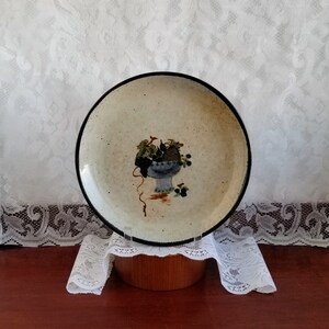 May include: A ceramic plate with a brown rim and a speckled beige surface. The plate features a hand-painted design of a blue and white vase filled with grapes and leaves.