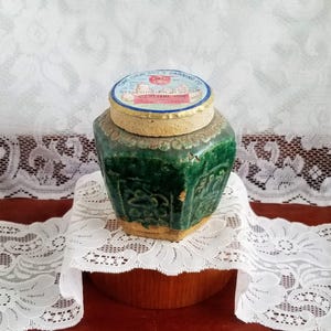 May include: A vintage, green ceramic jar with a lid. The jar has a textured surface and a distinctive shape. The lid features text on a blue and red label. The jar is placed on a white lace doily.