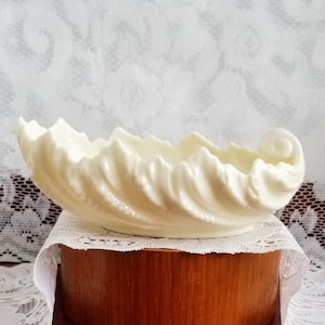 May include: Cream-coloured ceramic decorative bowl with a seashell design. The bowl has a wavy, textured exterior and a curled end. It sits on a wooden base covered with a white lace doily. The bowl is a decorative item.
