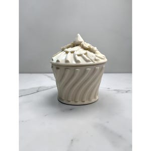 May include: A cream-coloured ceramic jar with a matching lid. The body features a vertical, swirling ribbed pattern. The lid is topped with a pointed finial and decorated with sculpted floral elements, including leaves and petals, in a matching cream tone.