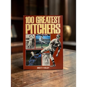 Op de afbeelding: Een hardcover boek getiteld 100 Greatest Pitchers door Brent P. Kelley. De donkerrode kaft bevat grote crèmekleurige tekst en vier afzonderlijke fotovakken met honkbalspelers die in verschillende werphoudingen op een veld staan afgebeeld.