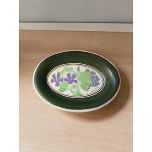 May include: An oval-shaped ceramic soap dish with a dark green rim and a white center. The center features a botanical illustration of purple mallow flowers with green leaves and the text MALVA printed in black lettering near the bottom edge.