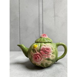 Puede incluir: Una tetera de cerámica verde con un diseño floral en relieve que presenta rosas rosadas y un pequeño capullo amarillo. La tetera incluye una tapa a juego con un remate en forma de rosa, un asa curva y un pico, todo con acabado brillante.