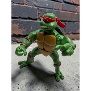 May include: A green plastic action figure of a turtle character wearing a red eye mask, a tan chest plate, and brown wrist and ankle bands. The figure is posed in a standing position with arms extended and clenched fists, showing a determined facial expression.