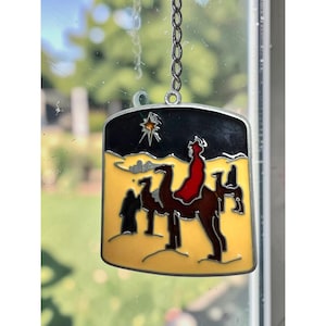 May include: A stained glass suncatcher featuring a desert scene with a camel and a figure in a red cloak under a black night sky. A bright yellow star shines above the landscape. The piece is framed in silver metal and hangs from a silver chain.