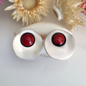 May include: A pair of silver-toned stud earrings with a circular design. Each earring features a vibrant red, speckled center stone set within a black border. The earrings are displayed on a white surface with dried flowers in the background.