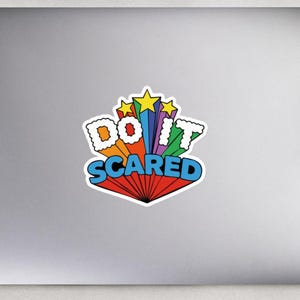 以下が含まれることがあります： 「DO IT SCARED」という言葉が大胆で遊び心のあるフォントで書かれたカラフルなステッカー。デザインは、星と赤、オレンジ、黄色、青、緑の鮮やかなカラーパレットを備えたスターバーストの背景が特徴です。