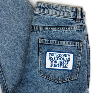 Peut inclure: Un jean en denim bleu avec un patch blanc cousu sur la poche arrière. Le patch a un texte bleu qui dit "You're only as cool as you treat people."