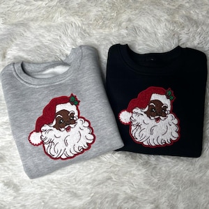 May include: Two sweatshirts with a black Santa Claus face embroidered on the front. One sweatshirt is gray and the other is black.