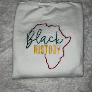 May include: White sweatshirt with an embroidered design of an outline of the continent of Africa in red and green. The words "Black History" are embroidered in yellow inside the outline.