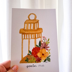 Könnte beinhalten: Ein Aquarellbild einer Laterne mit Blumen. Die Laterne ist hellbraun und hat ein Blumenmuster. Die Blumen sind in Rot-, Orange-, Gelb- und Lilatönen gehalten. Der Text "psalm 119:105" steht unter der Laterne.