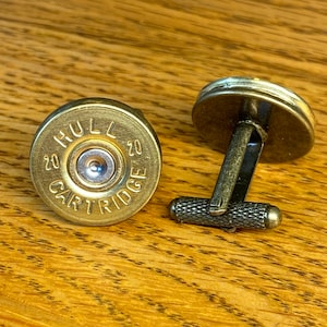 May include: A pair of gold-toned cufflinks featuring a 20 gauge shotgun shell casing with the words "HULL 20 CARTRIDGE 20" engraved on the front.