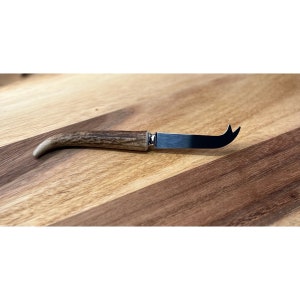 May include: A cheese knife with a curved blade and a handle made from a light brown antler. The blade is silver and the handle is attached with a silver bolster. The knife is designed for cutting cheese.