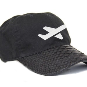 Może przedstawiać: Czarna czapka z daszkiem z białym logo samolotu. Daszek ma teksturowany wzór przypominający łuski. Czapka posiada regulowany pasek z tyłu. Stylowy dodatek.