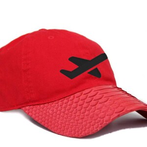 Może przedstawiać: Czerwona czapka bejsbolowa z czarnym motywem samolotu z przodu. Daszek ma teksturowany wzór przypominający łuski. Czapka jest w żywym czerwonym kolorze.