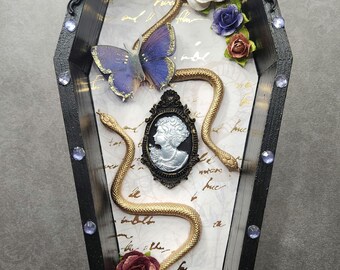 May include: Black coffin-shaped decorative box with ornate trim, embellished with rhinestones. Inside, a butterfly, golden snakes, a cameo, and paper roses in white, red, and blue adorn a patterned background. A unique gothic-inspired piece.