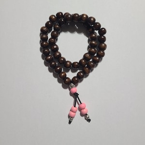 May include: A dark brown wooden bead necklace with a pink and silver tassel. The beads are round and evenly spaced. The tassel features pink beads and silver accents, suspended from a black cord.