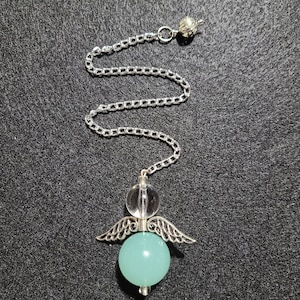 Puede incluir: Un péndulo con una cadena plateada y un diseño de ángel. El ángel tiene una cabeza de cristal transparente, alas plateadas y una esfera azul claro para el cuerpo. La cadena está unida a una cuenta plateada decorativa.