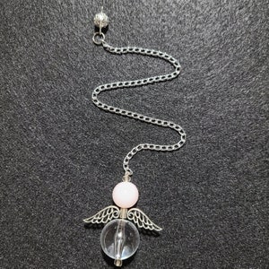 Op de afbeelding: Een zilverkleurige pendel met een ketting en een heldere kristallen bol met zilveren vleugels. Boven de kristallen bol bevindt zich een roze bol, en de ketting eindigt met een kleine zilveren bol. De pendel is ontworpen voor spirituele praktijken.