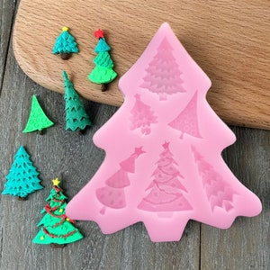 Op de afbeelding: Roze siliconen kerstboomvorm met 7 verschillende boomontwerpen. De vorm ligt op een houten oppervlak met verschillende gedecoreerde kerstboom.
