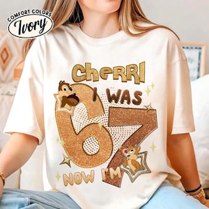 Peut inclure: T-shirt ivoire Comfort Colors avec l'inscription "Cherri I WAS 67 NOW I'M" en lettres marron et or pailleté. Le chiffre 67 est décoré de tamias et d'étoiles de dessins animés.