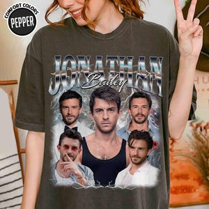 May include: Dark olive green t-shirt featuring a graphic of Jonathan Bailey with the name in a silver, vintage-style font. The design includes multiple images of the actor with a lightning bolt effect. The shirt has the Comfort Colors Pepper logo.