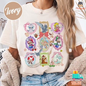 May include: Ivory-colored t-shirt featuring framed cartoon characters, including Mario, Luigi, Princess Peach, and Yoshi. The design incorporates decorative frames with pink and gold accents, creating a playful and nostalgic aesthetic.