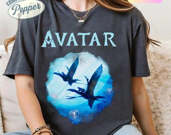 Pode incluir: Camiseta cinza escuro com a palavra "AVATAR" em azul claro, sobre um gráfico azul e branco de duas criaturas voadoras. A camiseta tem os logotipos "Comfort Colors" e "Pepper" no canto superior esquerdo.