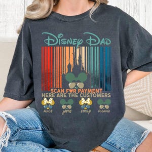 以下が含まれることがあります： ターコイズ色の「Disney Dad」の文字が入った濃いグレーのTシャツ。カラフルなバーコードのデザインで、お城のシルエットとミッキーマウス型のサングラスが特徴です。下には「Scan for Payment Here are the Customers」と書かれています。
