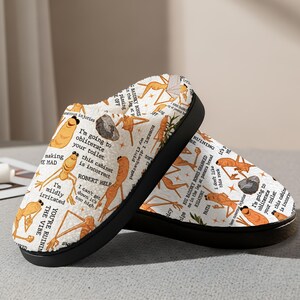 May include: Pair of white plush slippers with a black sole, featuring a repeating pattern of orange cartoon figures and text. The text includes phrases like "I'm going to obliterate your toilet" and "ROBERT HELP".