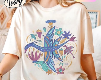 Pode incluir: Camiseta Comfort Colors marfim com um gráfico colorido de um pássaro estilizado com elementos florais e geométricos. O design inclui tons de azul, roxo, laranja e verde. A camisa tem o texto "Comfort Colors Ivory".
