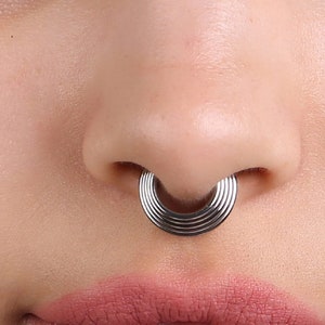 May include: A close-up of a person's nose with a silver septum ring. The ring is made of multiple thin bands that create a circular shape.