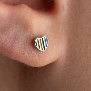 May include: A close-up of a person's ear with a small heart-shaped earring. The earring is silver with a rainbow stripe design.