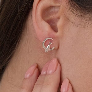 May include: Silver earrings featuring a crescent moon design with a small bird perched on top. The earrings are shown on a person's ear.