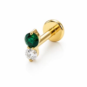 May include: A gold flat back labret stud featuring a vertical arrangement of two round-cut gemstones. The top stone is a deep emerald green, while the bottom stone is a clear, colorless white, both secured in a polished gold metal setting.