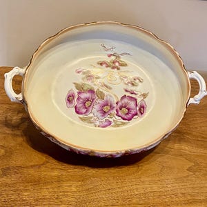 Puede incluir: Un cuenco antiguo de color crema con un borde festoneado y dos asas. El cuenco presenta un diseño floral con flores rosas y moradas, detalles dorados y un patrón delicado y ornamentado. El borde está adornado con oro.